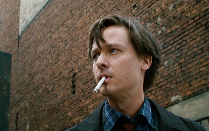 Ein junger Mann steht mit Zigarette im Mund vor einer hohen Mauer aus roten Ziegelsteinen