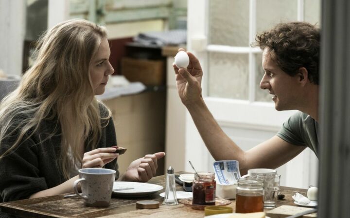 Eine blonde Frau und ein kurzhaariger Mann sitzen sich an einem Frühstückstisch gegenüber, er hält erklärend ein Ei in der Hand
